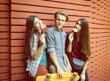 Two Beautiful Girls And Young Boy Have Fun Embracing And Licking Sweet Candy Outdoor Against Red Brick Wall