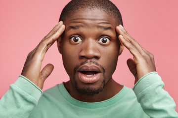 Wall Mural - Close up portrait of bug eyed stupefied black man keeps hands on head, stares at camera, realizes that he forgot to pay bills, poses against pink studio background. Emotions and lifestyle concept