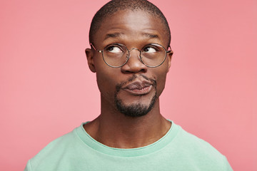 Indoor shot of handsome dark skinned male curves lips, looks doubtfully aside, has hesitations, thinks what to do or makes planes for future, has dreamy pensive expression. Uncertainty concept