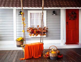 interior of an autumn patio. swing is adorned with autumn leaves and orange knitted plaid. Basket with pumpkins and autumn vegetables. The window is decorated with autumn decor.