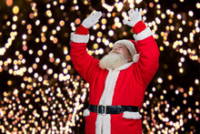 Santa Claus On New Year Lights Background. Santa Claus Outdoors On Garlands Background With Raised Hands And Looking Upwards. Santa Claus Making A Magic.