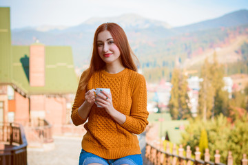 Cute and nice young woman drinking coffee at terrace on a sunny morning in mountain. Pretty lady in stylish casual yellow sweater and shorts. Warm soft cozy image, breakfast on the balcony  