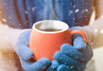 A cup of hot drink, tea or coffee, in winter mittens. Snow falls. New Year concept. Christmas mood.