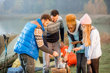 Multi ethnic group of friends dressed in sweaters having a dinner eating fondue during the outdoor recreation at the camping in the evening