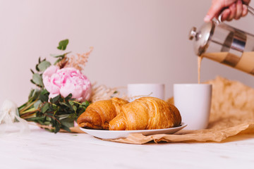 Delicious healthy breakfast with two fresh flaky croissants served on a plate with two cups of coffee and a bouquet of summer wildflowers on kitchen table. Woman's hand pouring coffee