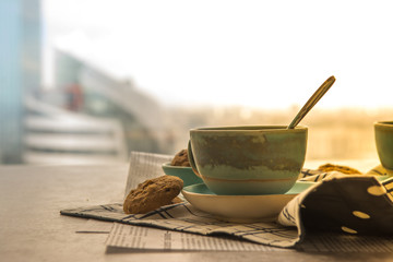 Cup of black coffee with spoon on supported dish on fabric on black and white newspaper is put on hard table in the early morning with bright yellow light and some biscuits.