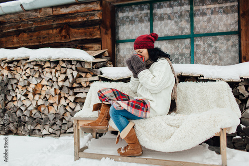 Plakat Kobieta ubrana ciepłe siedzenie na zewnątrz domku na chłodne zimowe wieczory. Portret kobieta cieszy się jej filiżankę herbata na zewnątrz dom na wsi w zimie.