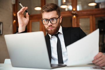 Poster - Young concentrated bearded businessman in formalwear looking at laptop screen holding papers while sitting at workplace