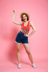 Poster - Full length portrait of happy playful woman in summer wear and straw hat, pointing with finger upward, looking at camera