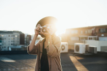 Trendy and attractive beautiful woman with analog vintage film camera makes photos on rooftop at sunset time, creates content for social media channels and applications, popular influencer