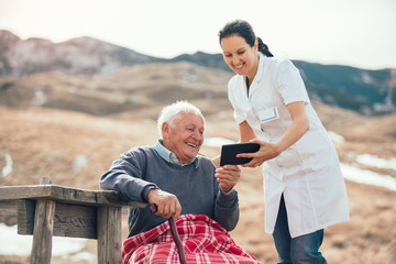 Wall Mural - Smiling caregiver nurse and  disabled senior patient using digital tablet outdoor