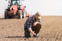 Young Farmer Examing Dirt While Tractor Is Plowing Field