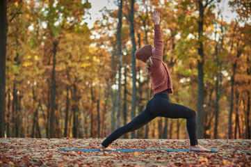 Beautiful young woman practices yoga asana Virabhadrasana 1 - warrior pose on the wooden deck in the autumn park.