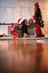 Wall Mural - Charming happy couple with Santa hats lying on a carpet under a red blanket while girl trying to kiss her boyfriend in the living room for Christmas holidays.