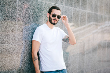 Wall Mural - Young man wearing white blank t-shirt with beard in glasses, standing on the street on city background. Street photo