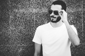 Wall Mural - Young man wearing white blank t-shirt with beard in glasses, standing on the street on city background. Street photo