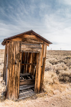Old Wooden Outhouse In A Desert. Sage Brush Is In The Background. A Blue Sky With Wispy Clouds.