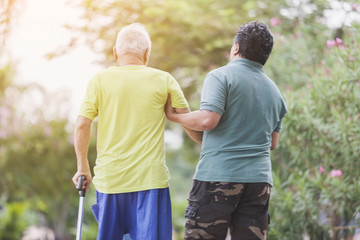 rear view - portrait of Physiotherapist or male nurse helping or holding hands an elderly gentleman in park. the mature man walking exercise together. Professional assistant supporting elderly man.