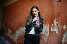 Stylish Brunette Girl Wear On Leather Jacket And Shorts With Mobile Phone At Hands Against Orange Wall.