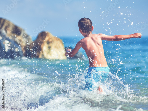 Plakat Cute Kaukaski chłopiec biegnie w wodzie wzdłuż brzegu morza. Widok z tyłu.