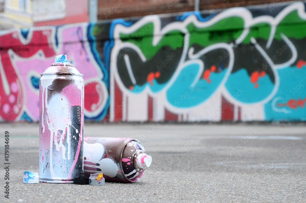 Several used spray cans with pink and white paint and caps for spraying paint under pressure is lies on the asphalt near the painted wall in colored graffiti drawings - obrazy, fototapety, plakaty 