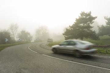 Foggy asphalt curved road with cars passing through the forest. Weather with low visibility in the region of Normandy, France. Country landscape on misty day. Toned