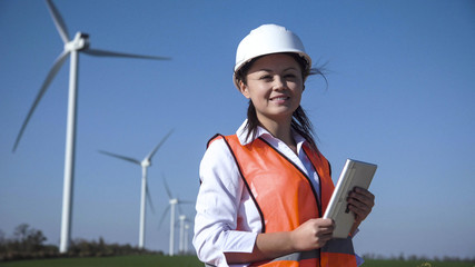 Cheerful woman wearing hard hat standing against turbines at wind farm on sunny day