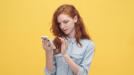 Wall Mural - happy surprised ginger woman in denim shirt using her smartphone over yellow background