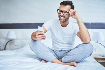 Wall Mural - Man sitting on bed and using phone smiling