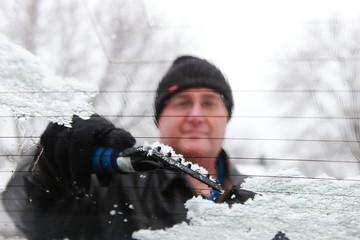 Man scraping snow and ice from car window , shot through wet windshield,