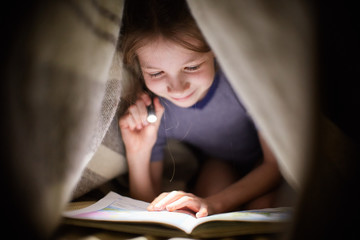 little girl is reading a book under a blanket with a flashlight in a dark room at night