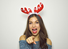 Happy Young Woman With Reindeer Horns On Her Head Looks And Pointing At Camera On Gray Background. Christmas Woman.