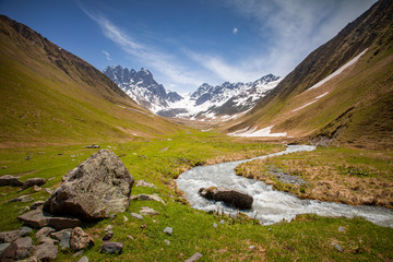 Wall Mural - Summer landscape with river and mountain snowy peak, Kazbegi national park, Georgia. The main Caucasian ridge. Green hills and small mountain river in Caucasus foothills, above view