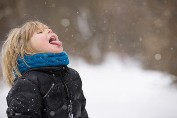 Cute blond kid boy catching snowflakes with his tongue. Walking in a winter park. Child having fun with snow outdoors.