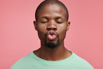 Wall Mural - Horizontal portrait of dark skinned man roundes lips as going to kiss girlfriend, closes eyes with pleasure, poses in pink studio. Bearded African American young male makes grimace, stands alone