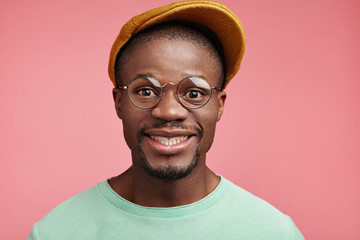 Wall Mural - Glad smiling hipster male wears fashionable cap, glasses, has good mood as spends free time in merry company, expresses positive emotions. Fashionable dark skinned guy poses against pink wall