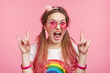 Crazy mad excited female model gestures, shows cool sign, opens mouth widely, poses against pink studio background. Astonished funny young woman coquette screams loudly, wears fashionable clothes