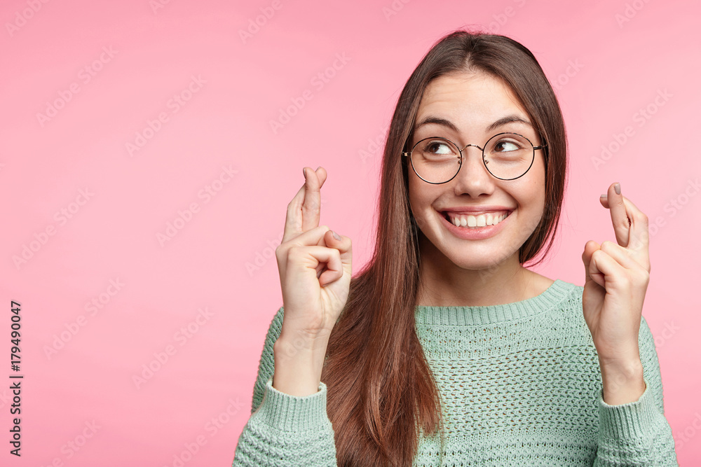 Indoor shot of glad brunette woman wears spectacles, smiles broadly, keeps fingers crossed, hope for good luck during long trip, isolated over pink background with copy space for your text or hearder - obrazy, fototapety, plakaty 