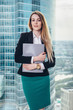 © undrey - Young female manager standing in the office holding a folder in hands against window with a view on city business center