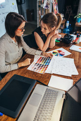 Wall Mural - young female graphic designers sitting at desk with laptop, color charts and some sketches discussin