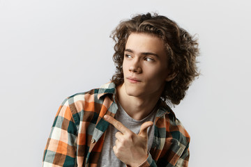 Wall Mural - Check this out. Close up portrait of cool fashionable young man with curly hair pointing index finger sideways, indicating copy space on blank studio wall for your text or advertising content