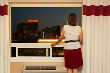horizontal image of a caucasian woman wearing a red skirt and white shirt standing by a big window in her hotel room looking out at the skyline of the city at night time.