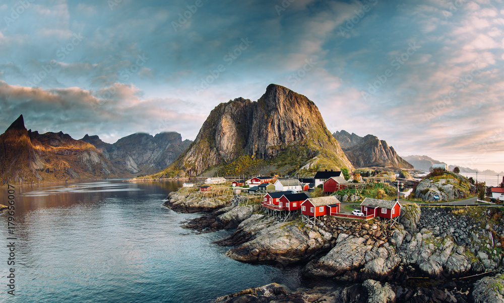 Norwegian fishing village at the Lofoten Islands in Norway. Dramatic sunset clouds moving over steep mountain peaks - obrazy, fototapety, plakaty 