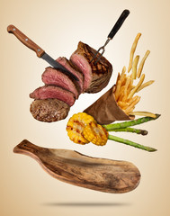 Poster - Flying beef steaks with grilled vegetable and french fries served on wooden cutting board.