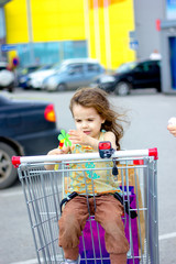 Sticker - Little kid girl sitting in a shopping cart with the shopping bags inside.