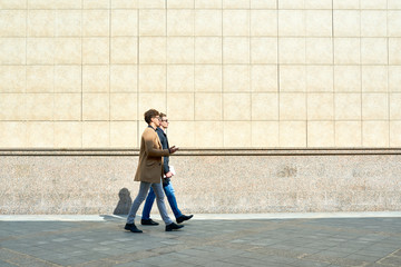 Side view portrait of two modern handsome men wearing autumn coats walking in city street against beige concrete wall