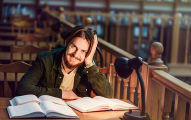 Portrait of smiling young bearded student man reading in a library hall on table with lot of books and lamp, indoor dusk time, education concept