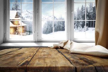 Wall Mural - Wooden table with free space for your product or promotional text. Open window with snowfall. Landscape of winter forest mountains and small dock at the end of the village.