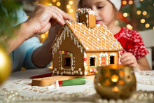 Father And Adorable Daughter In Red Hat Building Gingerbread House Together. Beautiful Decorated Room With Lights And Christmas Tree, Table With Candles And Lanterns. Happy Family Celebrating Holiday.