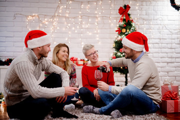 Wall Mural - Group of young attractive friends pouring and drinking a red wine while sitting on a carpet at home for Christmas holidays.